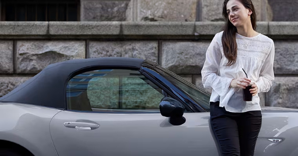 Woman standing beside a convertible car holding a beverage.